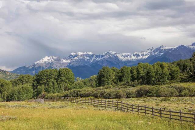 Snow-dusted peaks of the Sneffels Range rising above green ranchland and aspen groves behind a split-rail fence