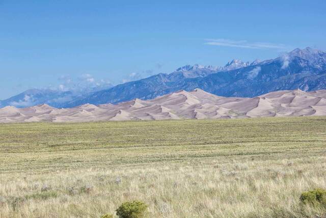 Tall sand dunes rising against a backdrop of jagged, cloud-wrapped mountains beyond a grassy valley floor