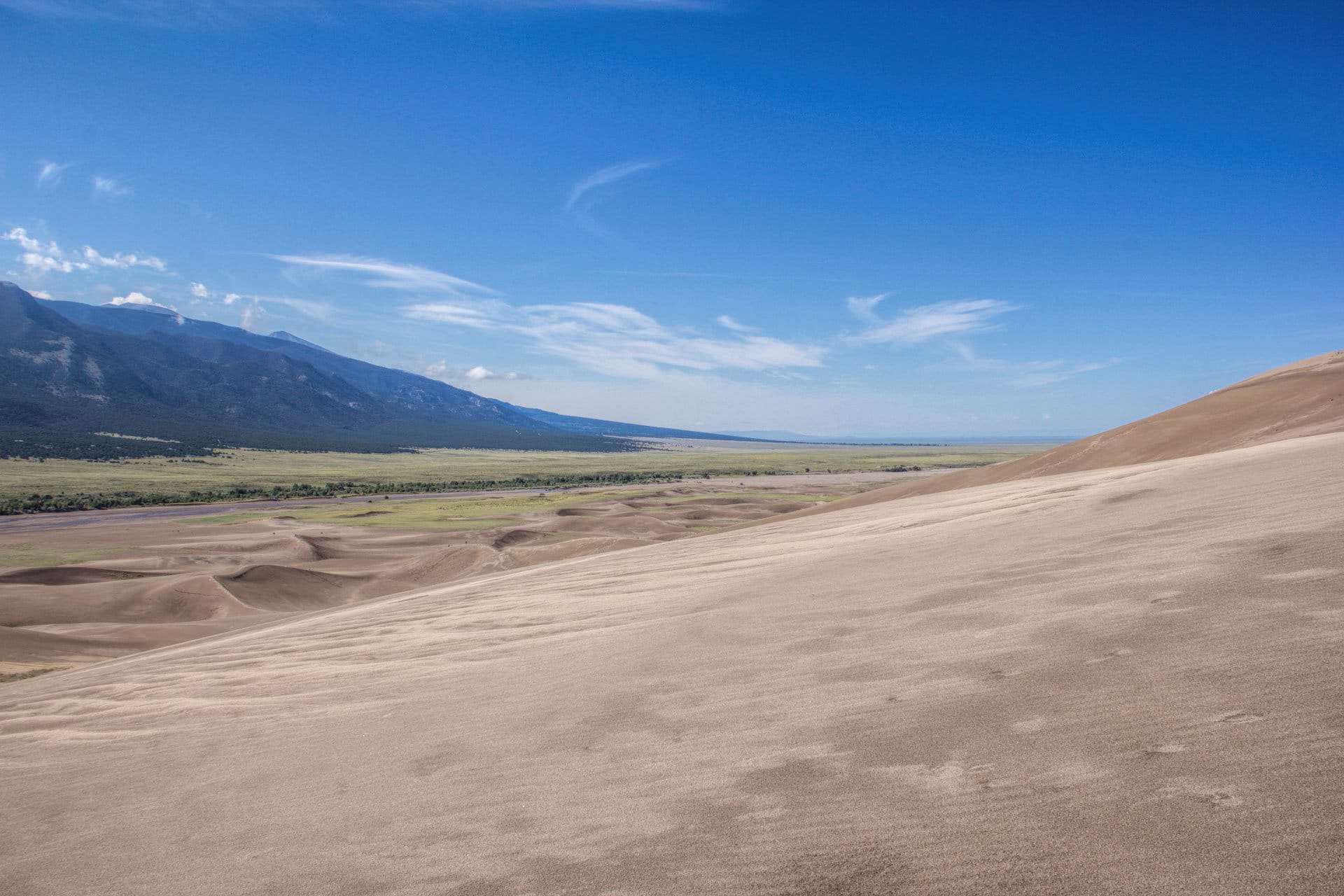 A high dune overlook across a rolling sea of sand toward a green valley and distant mountains under wispy clouds