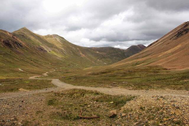 A rocky four-wheel-drive road winding through a wide alpine valley beneath layered, cloud-covered peaks