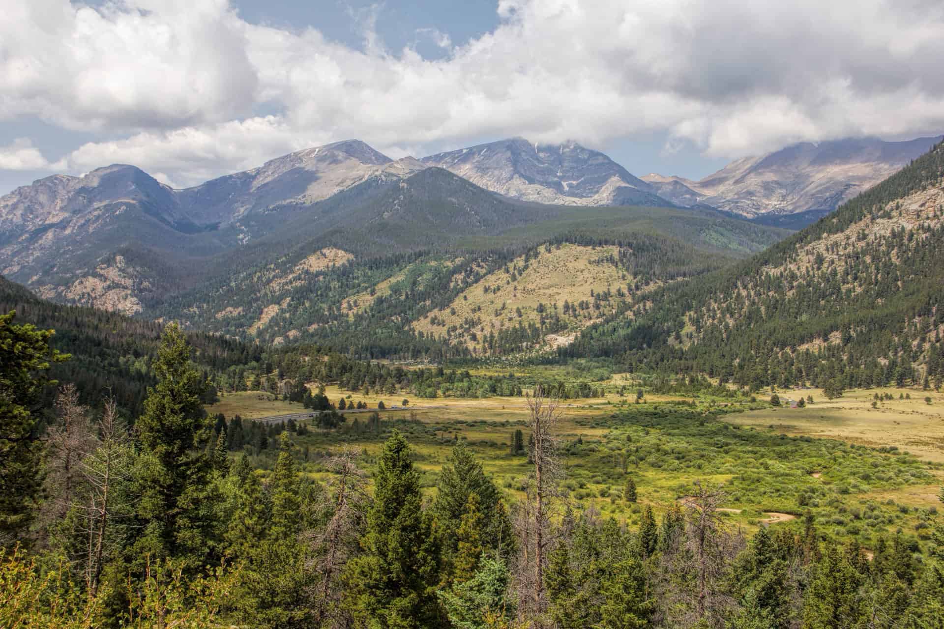 A forested valley in Rocky Mountain National Park with a road winding below rugged, cloud-capped peaks