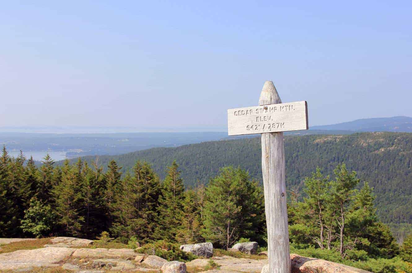 A wooden trail sign for "Cedar Swamp Mtn." overlooking a forested valley and distant bay