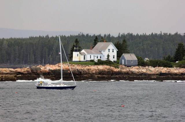 A lighthouse keeper's house on a rocky point along the Maine coast, with a sailboat passing by
