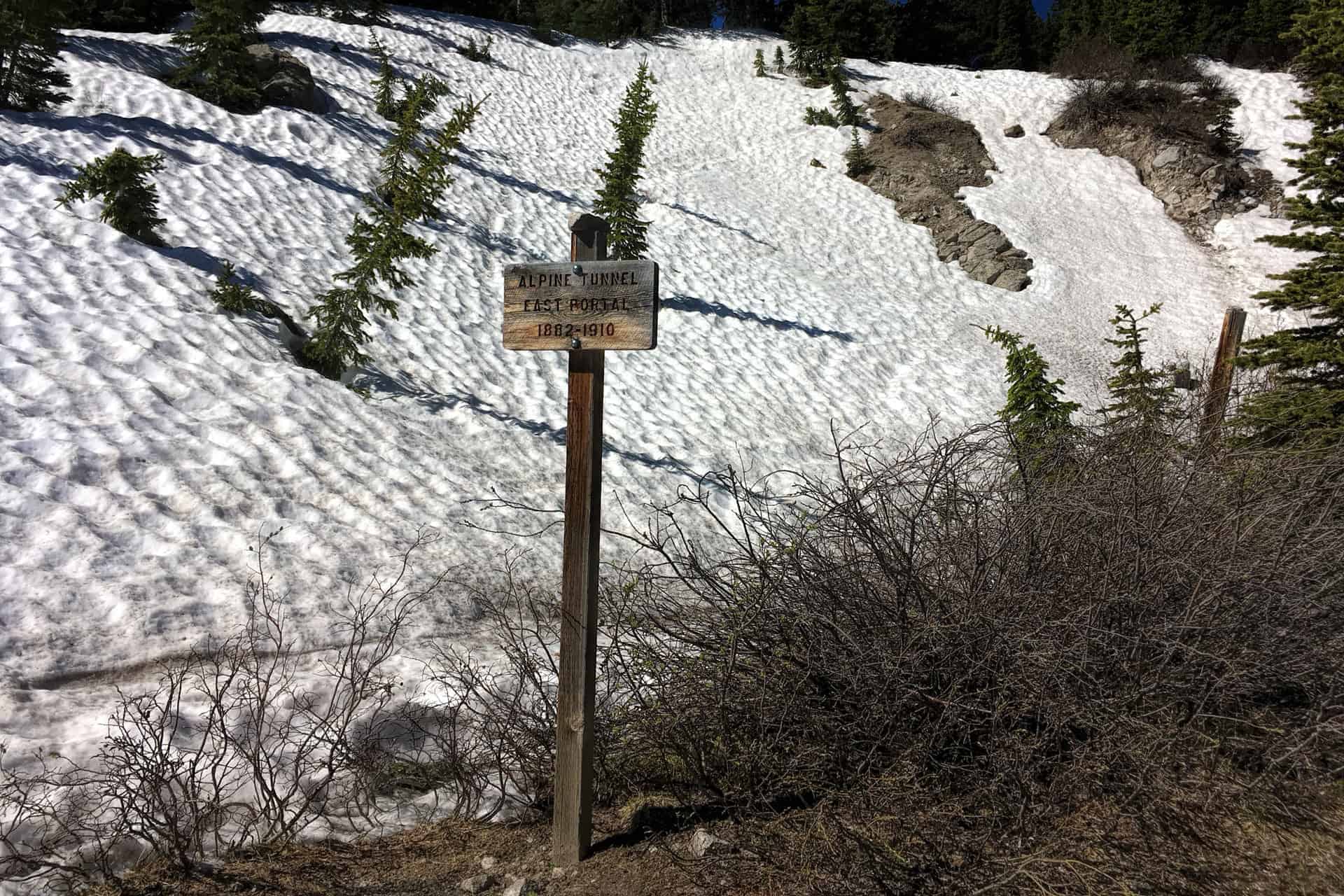 A weathered wooden trail sign reading 'Alpine Tunnel East Portal 1882-1910' standing before a wide snowfield