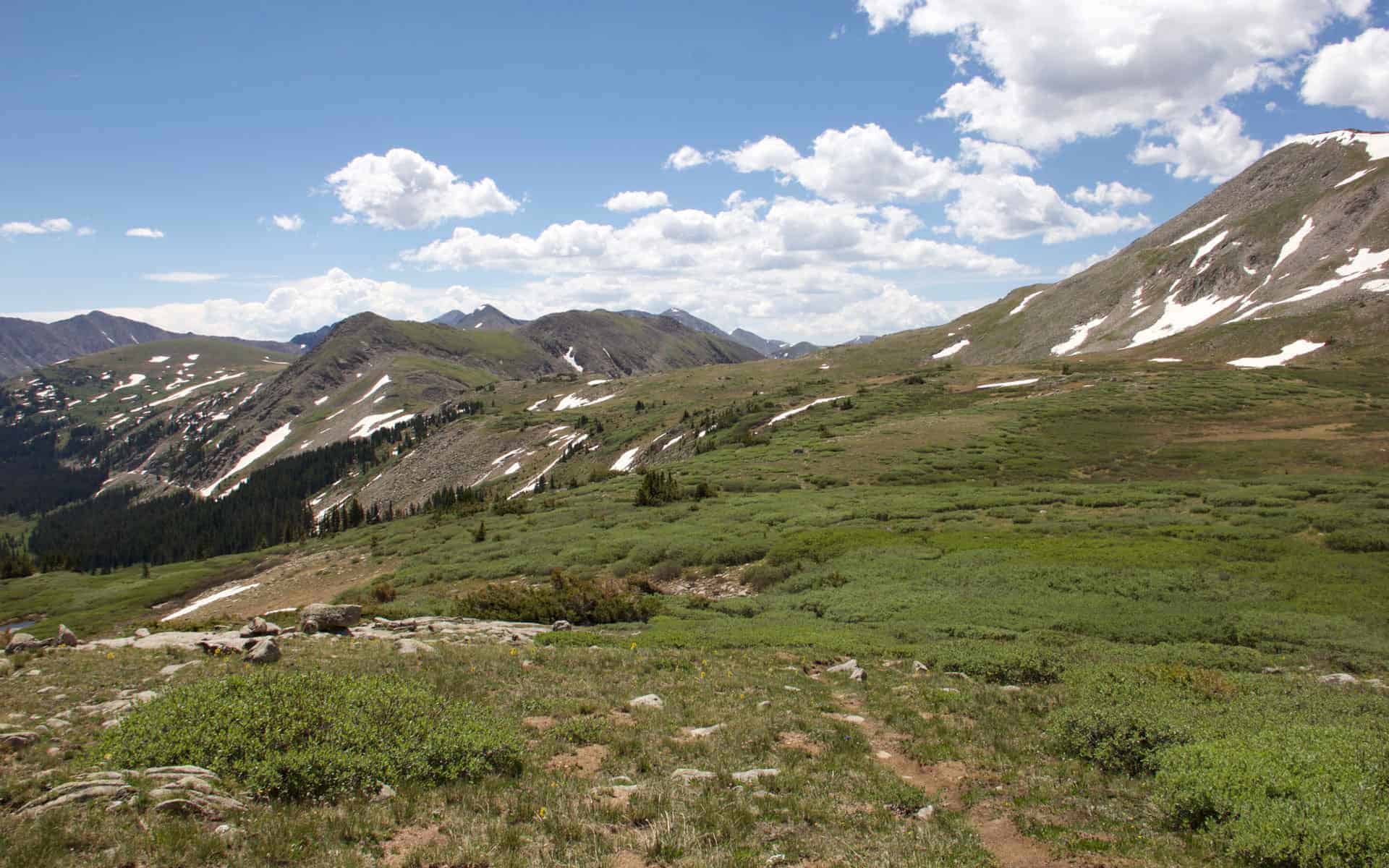 A wide mountain valley view with scattered snow patches, layered peaks, and clouds gathering on the horizon
