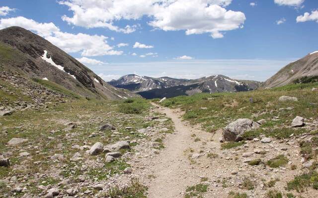 A narrow dirt trail winding through a rocky alpine basin toward distant snow-streaked mountains under a partly cloudy sky
