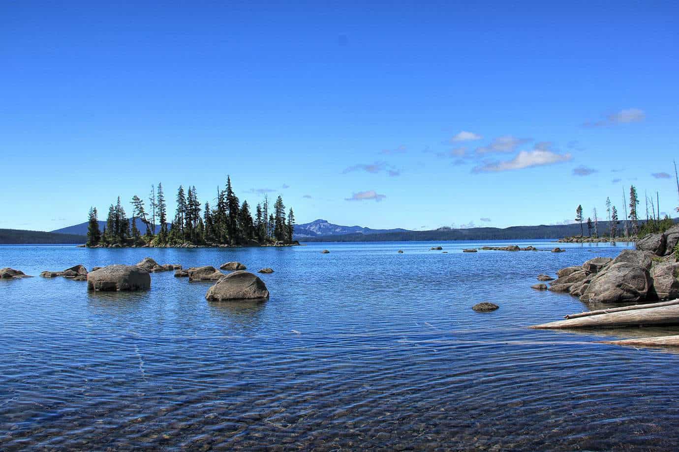 A rocky lake shoreline with a small forested island and mountains in the distance under a blue sky