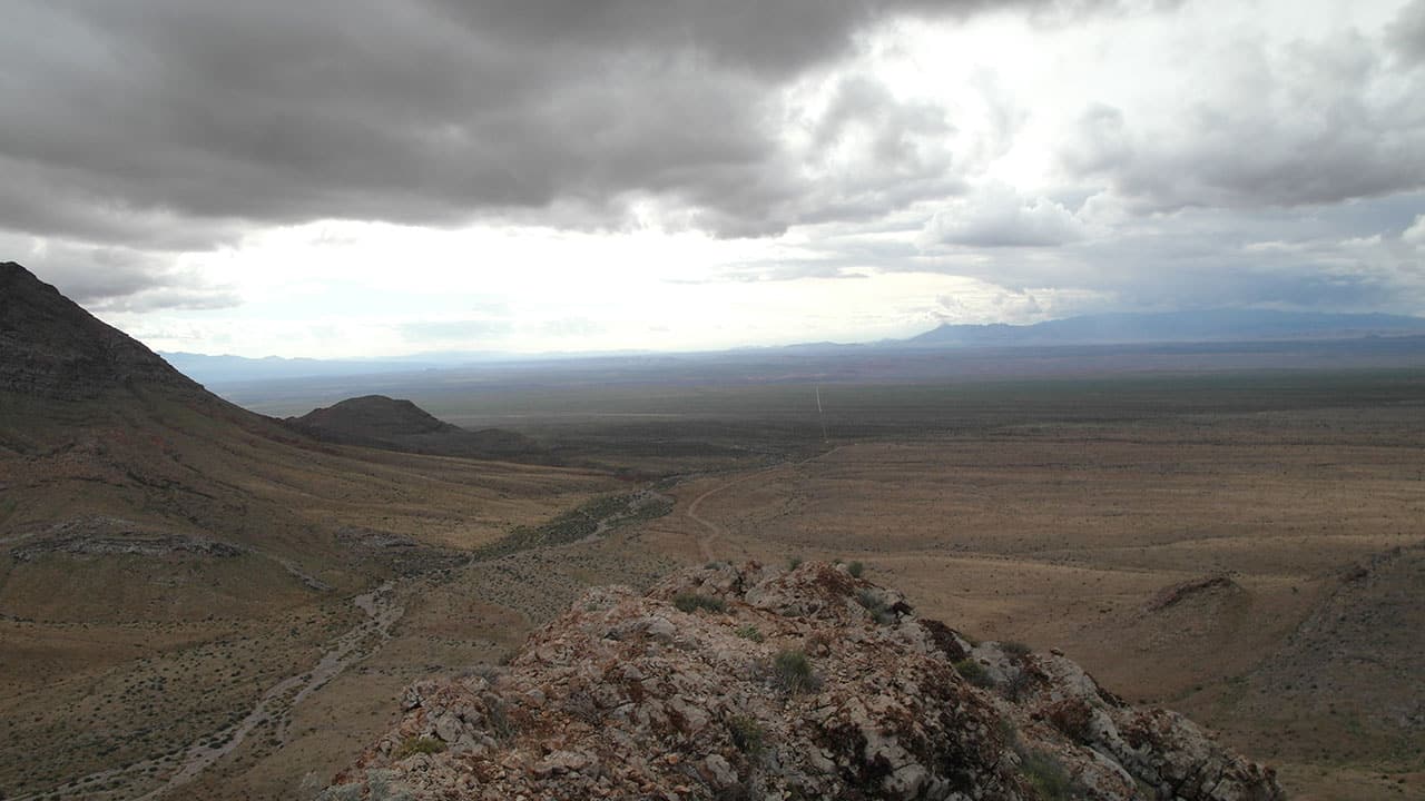 A wide desert valley view beneath dramatic dark storm clouds, with a dirt road winding below