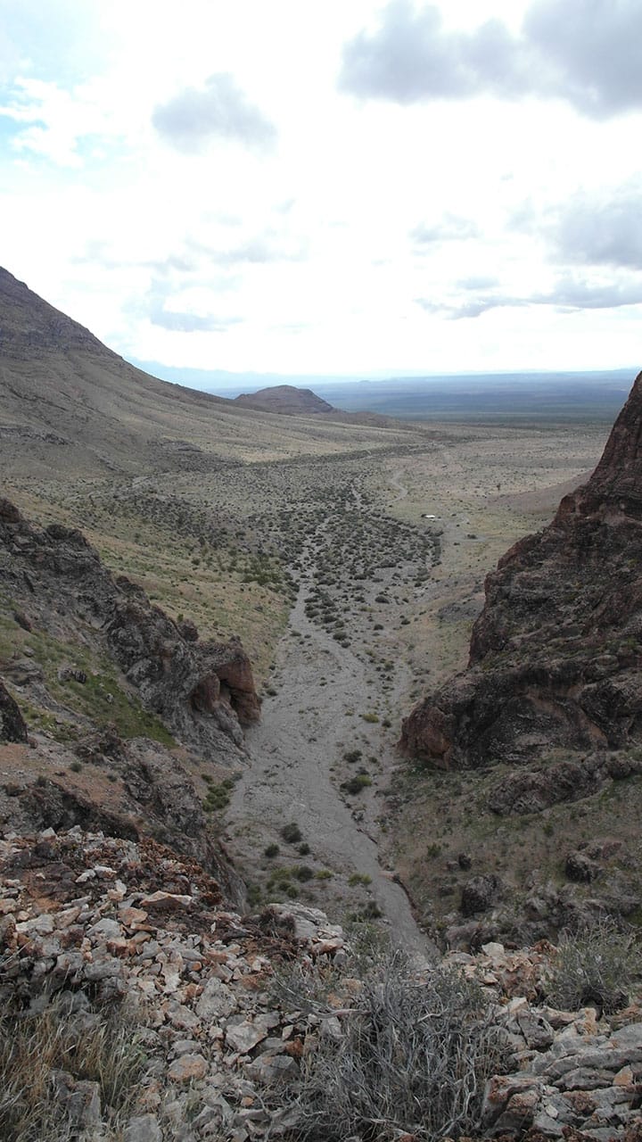 A vertical view down a rocky wash between canyon walls opening onto a wide cloudy valley