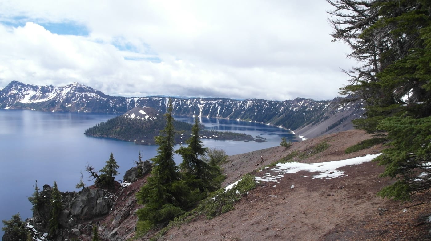 A view down a snow-dusted slope to Crater Lake and Wizard Island, framed by evergreens
