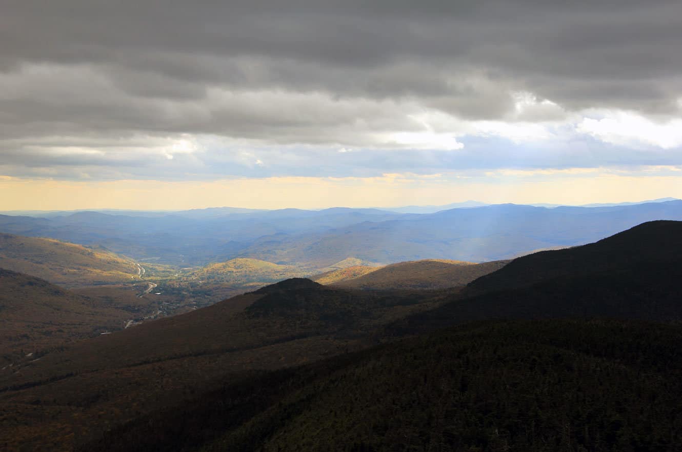 Sunlight breaking through clouds over a valley of fall-colored hillsides and a distant river