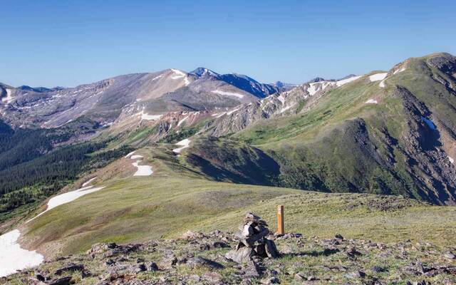 A wooden trail marker atop a rock cairn on a grassy ridge, with layered snow-patched peaks stretching into the distance