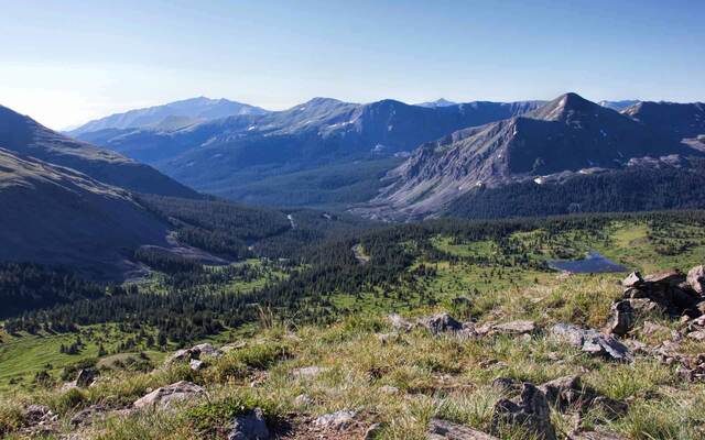 A wide view down a forested valley toward layered mountain ridges, with a small lake visible far below