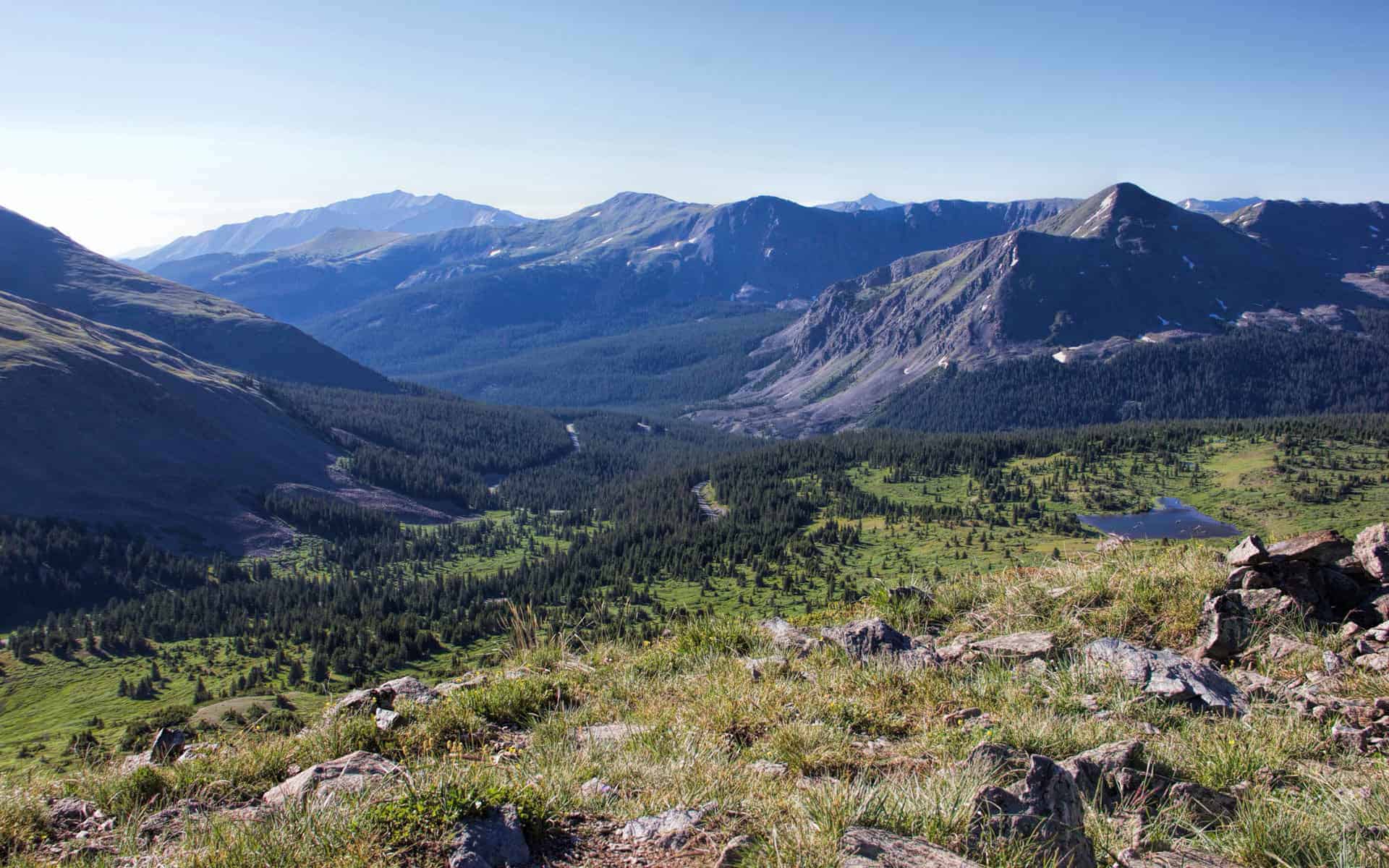 A wide view down a forested valley toward layered mountain ridges, with a small lake visible far below