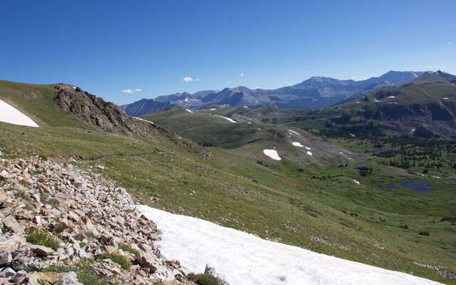 A rocky ridge trail with patches of snow, overlooking rolling green hills and distant peaks
