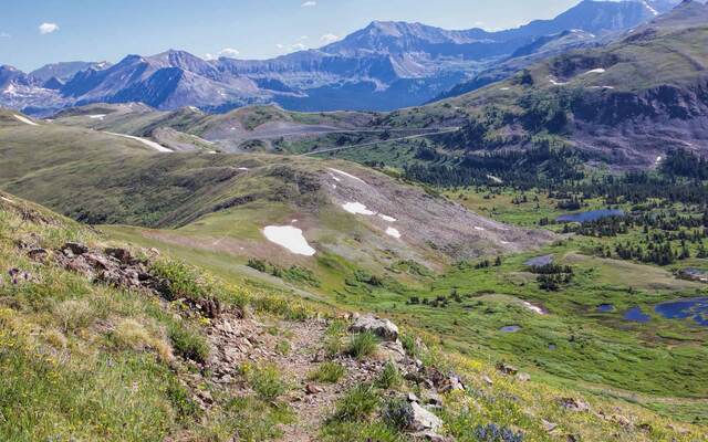 A wildflower-lined ridge trail descending toward a green valley dotted with small ponds, framed by distant mountains