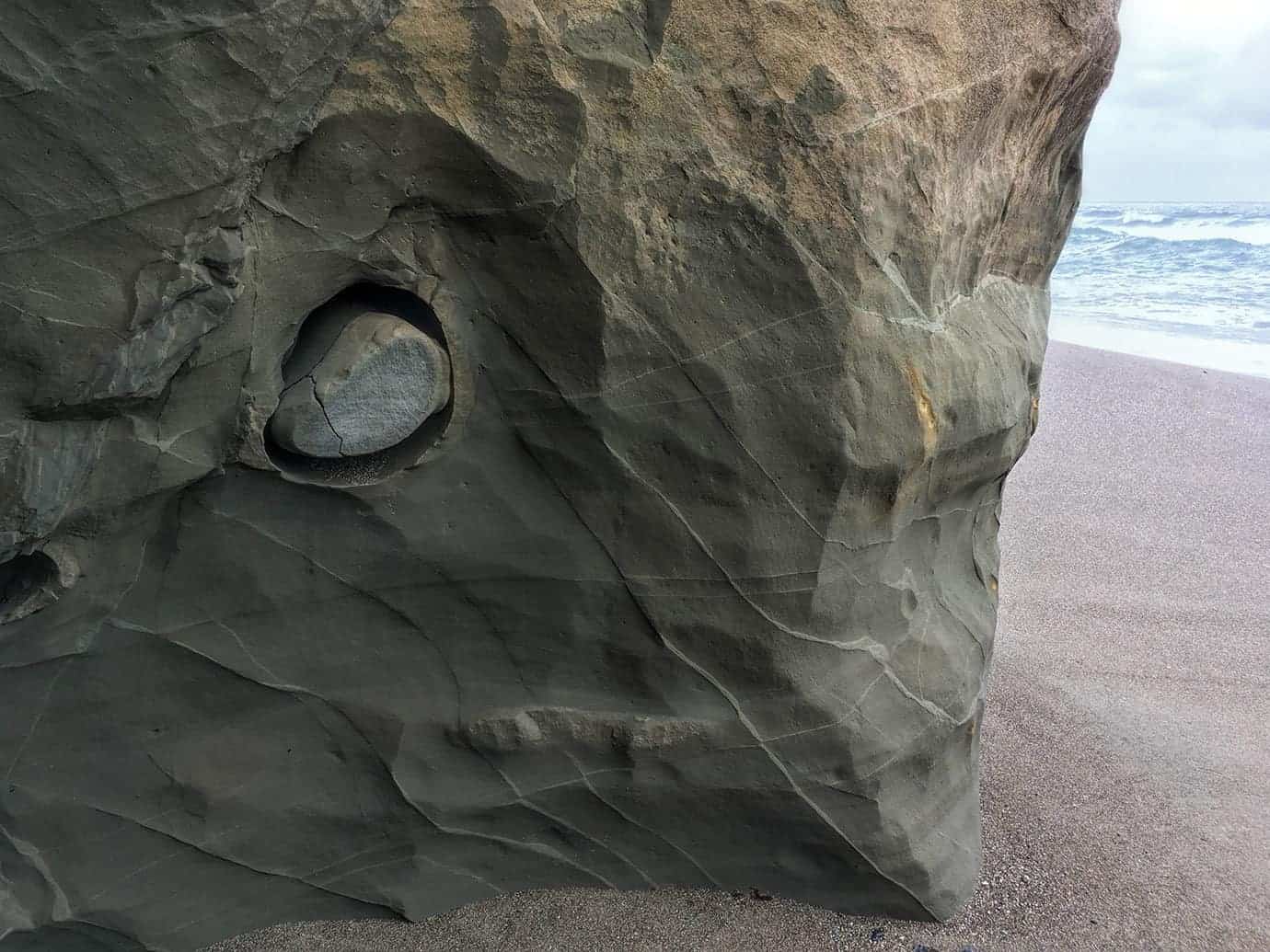 A round stone embedded in a hollow of a weathered sandstone rock face, with the ocean and surf visible beyond