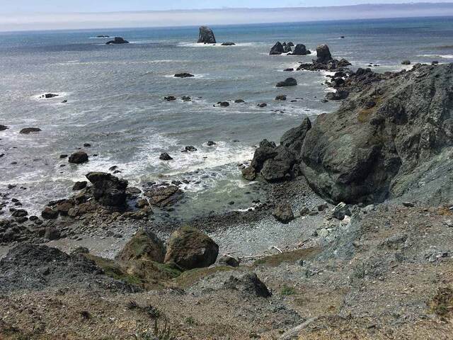 A wide view from a cliff edge across the ocean toward numerous small sea stacks scattered along the hazy coastline