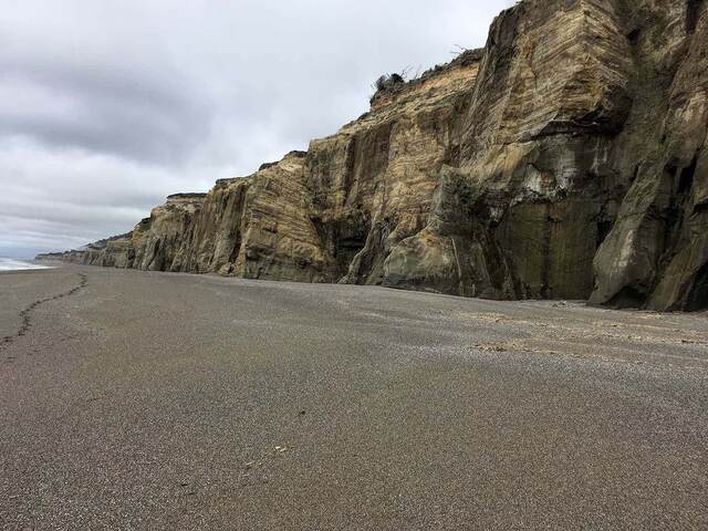 Layered sandstone cliffs rising above an empty beach under a cloudy sky, waves lapping at the base