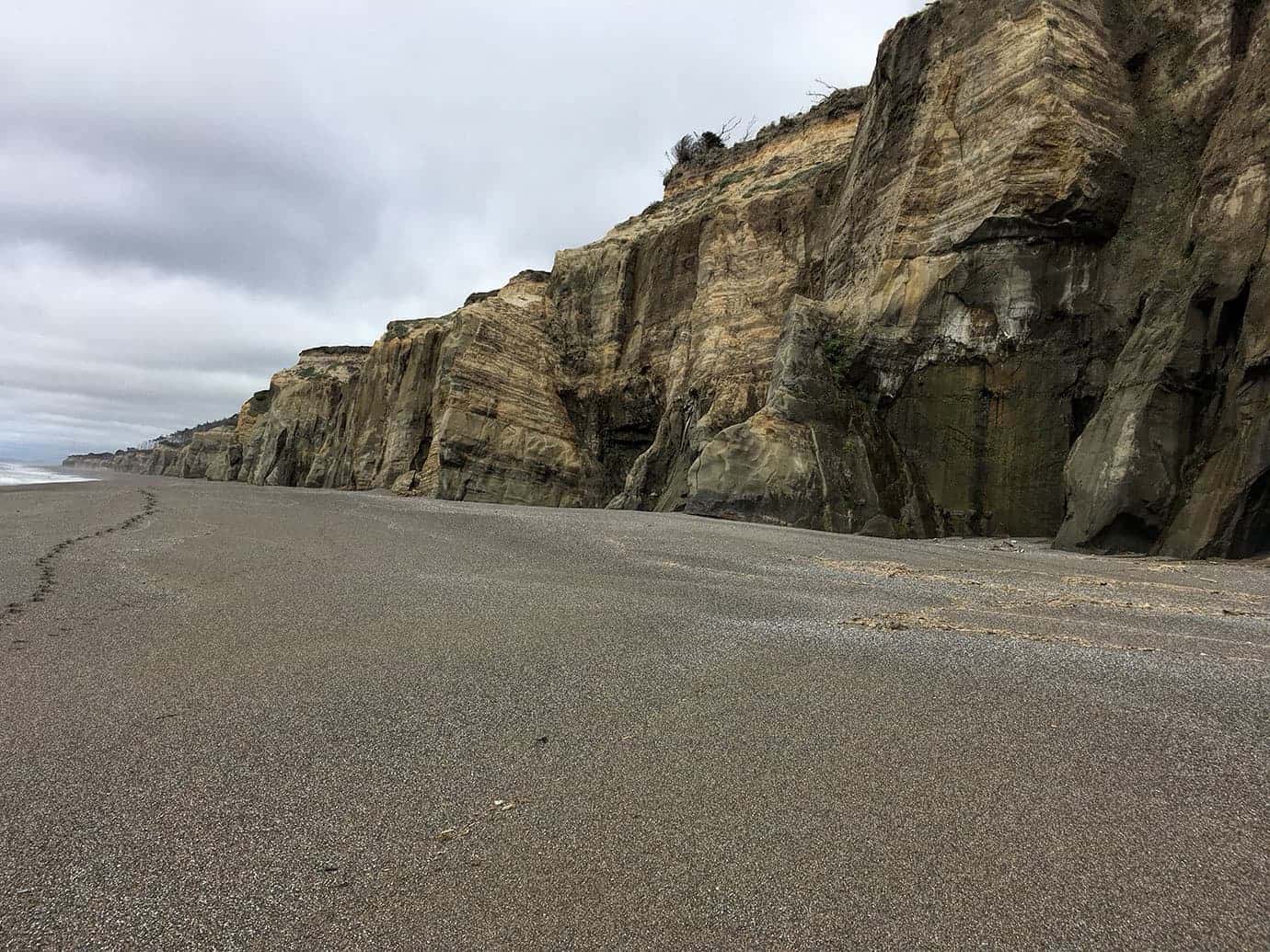 Layered sandstone cliffs rising above an empty beach under a cloudy sky, waves lapping at the base
