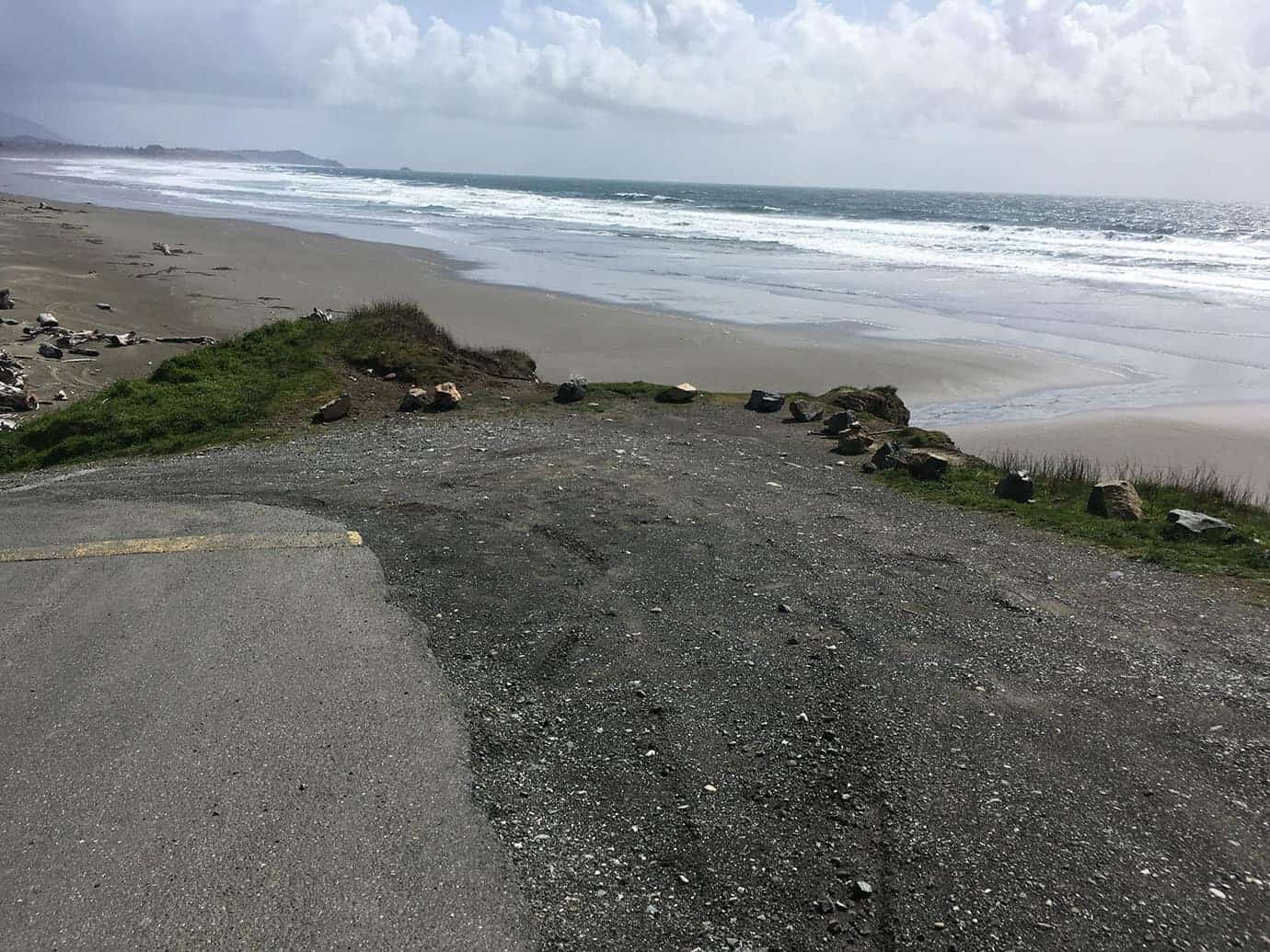 A paved beach access path leading down to the ocean, framed by dune grass and dark volcanic rocks