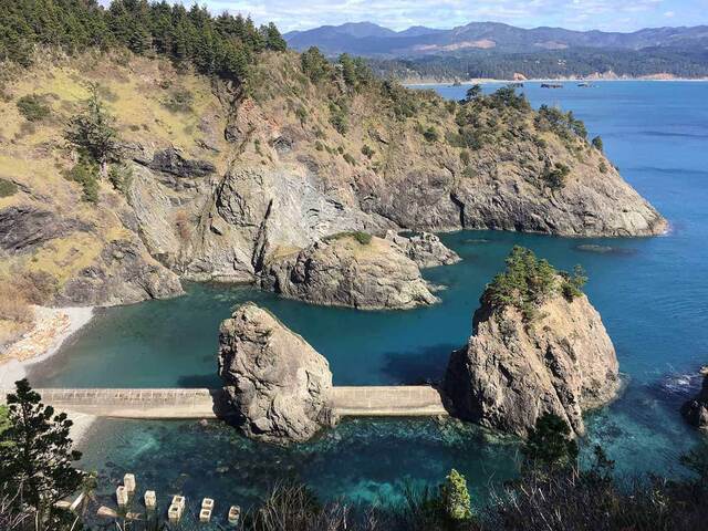 An aerial view of a rocky cove with the remains of an old boat ramp crossing between two sea stacks, turquoise water below and mountains beyond