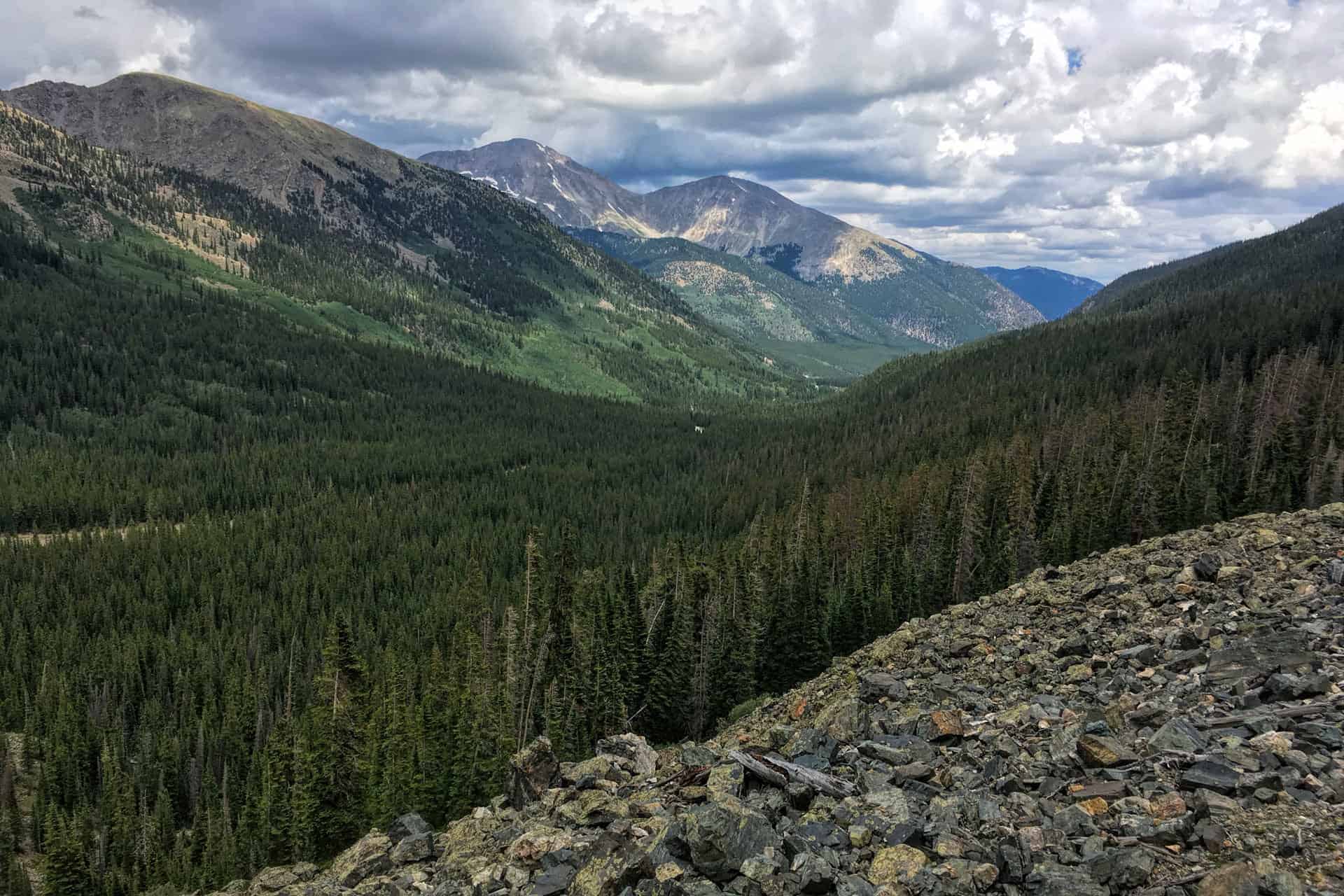 A view down a forested valley toward layered mountain peaks under a partly cloudy sky