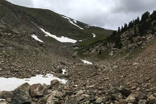 A rocky alpine gully streaked with patches of snow, descending toward a green valley and distant mountains under a cloudy sky