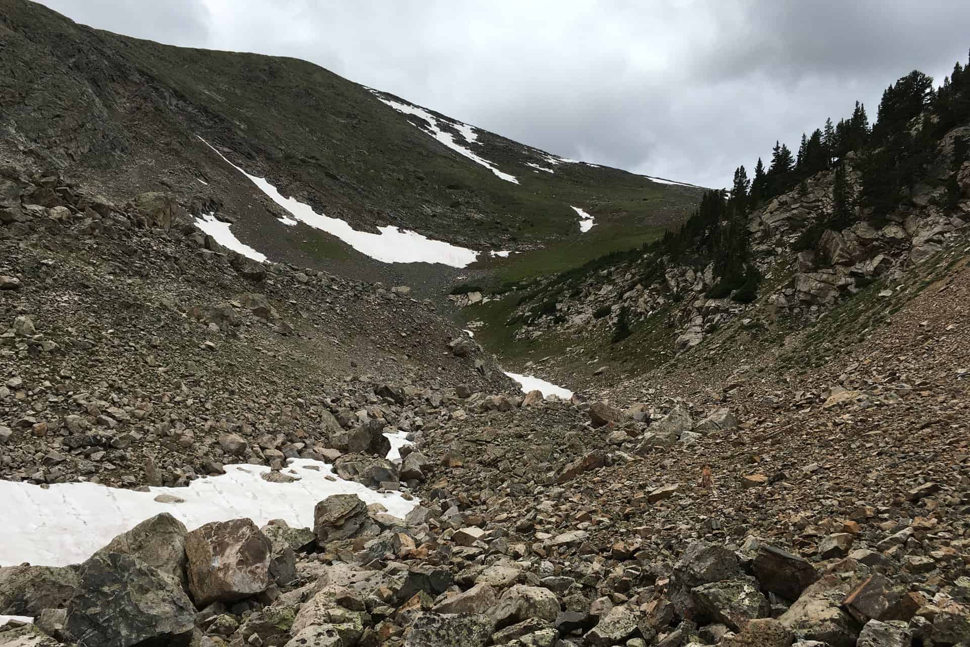 A rocky alpine gully streaked with patches of snow, descending toward a green valley and distant mountains under a cloudy sky
