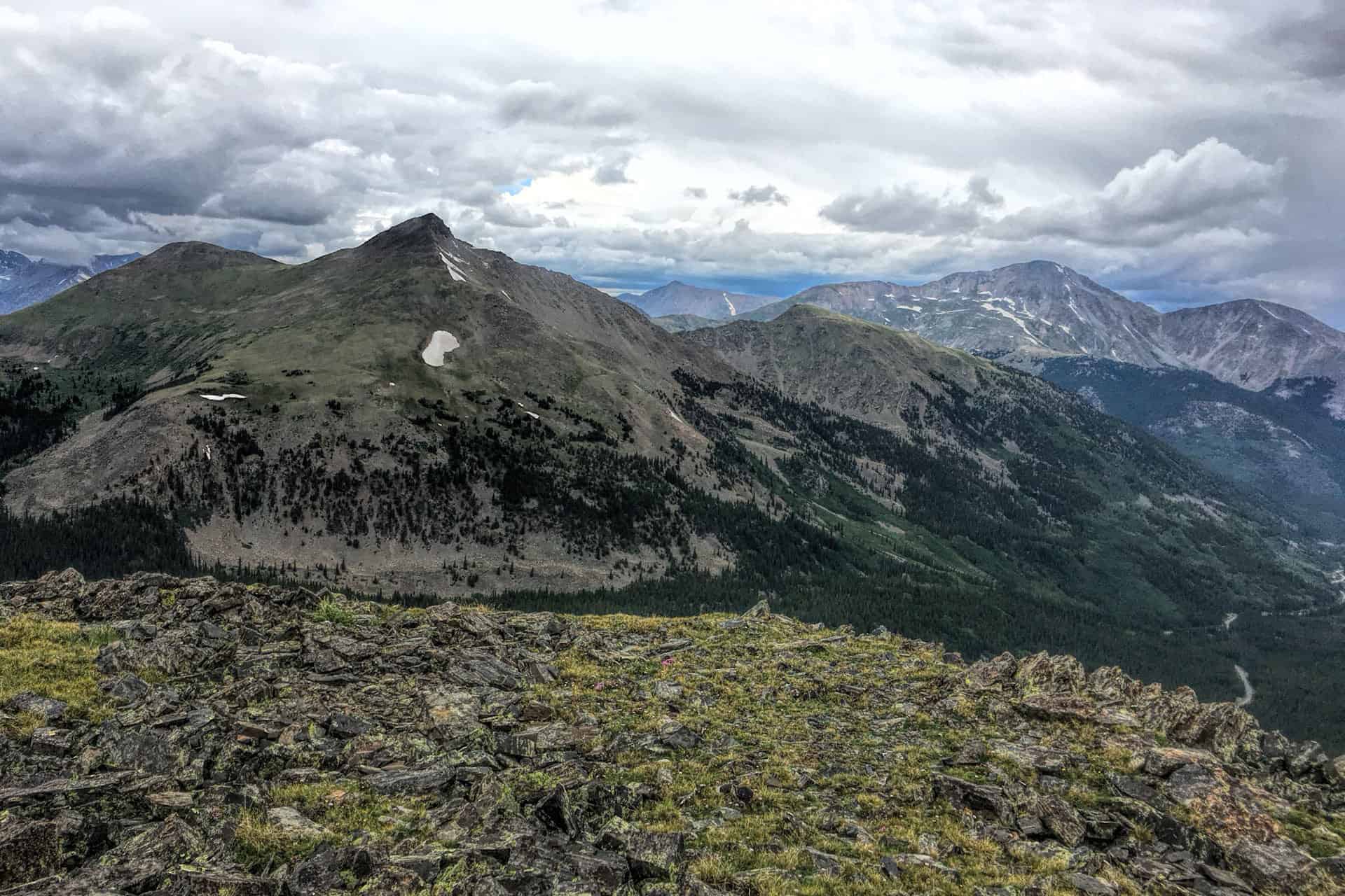 A grassy, rocky summit ridge with a view of a sharp peak and rolling mountains streaked with patches of snow