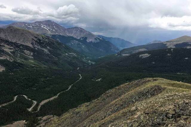 A forested valley with a winding road following a stream, mountains rising on either side and a rain shower visible in the distance