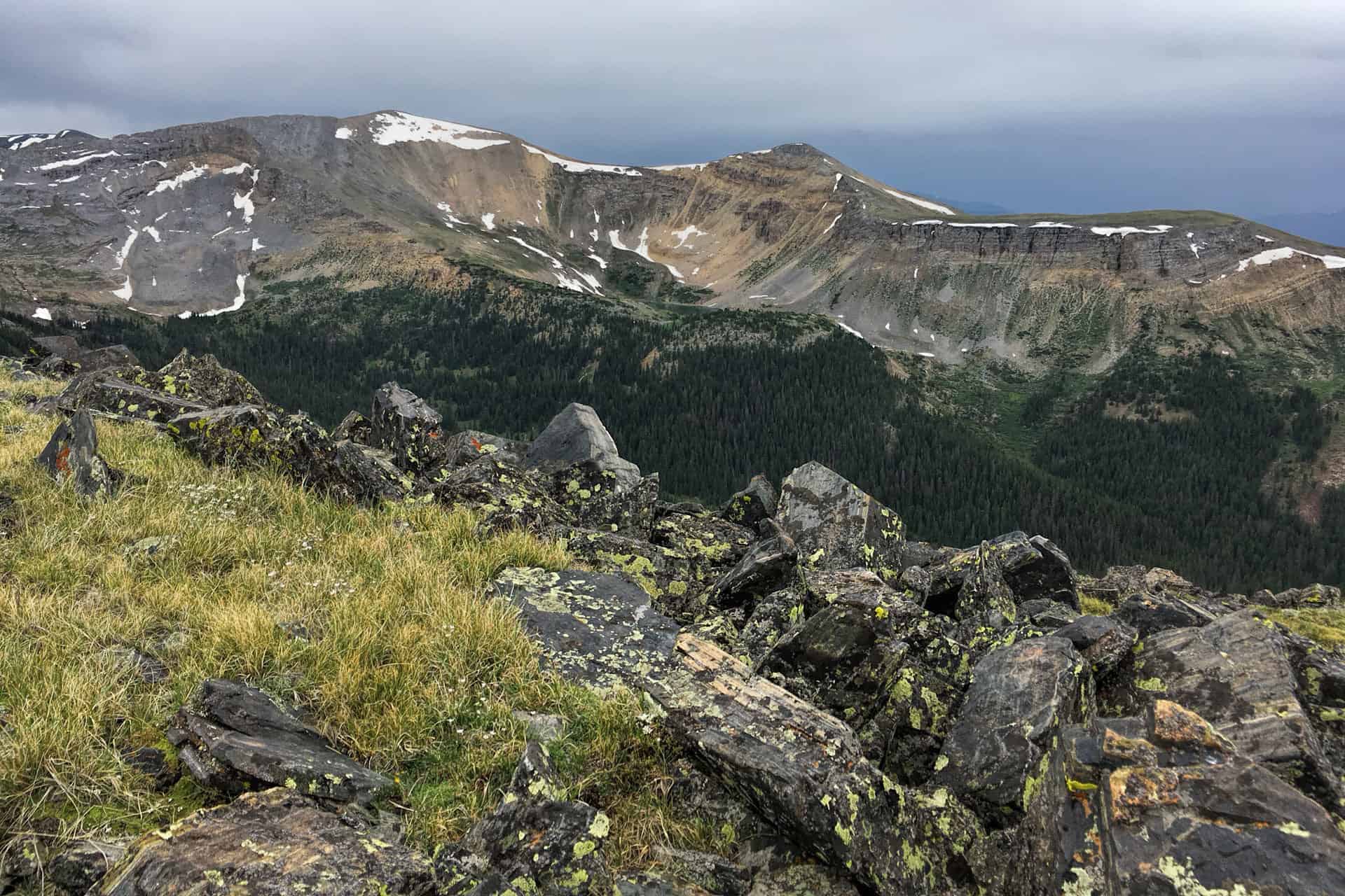 A rugged alpine cirque with snow-streaked cliffs and slopes, seen across a grassy, rock-strewn ridge