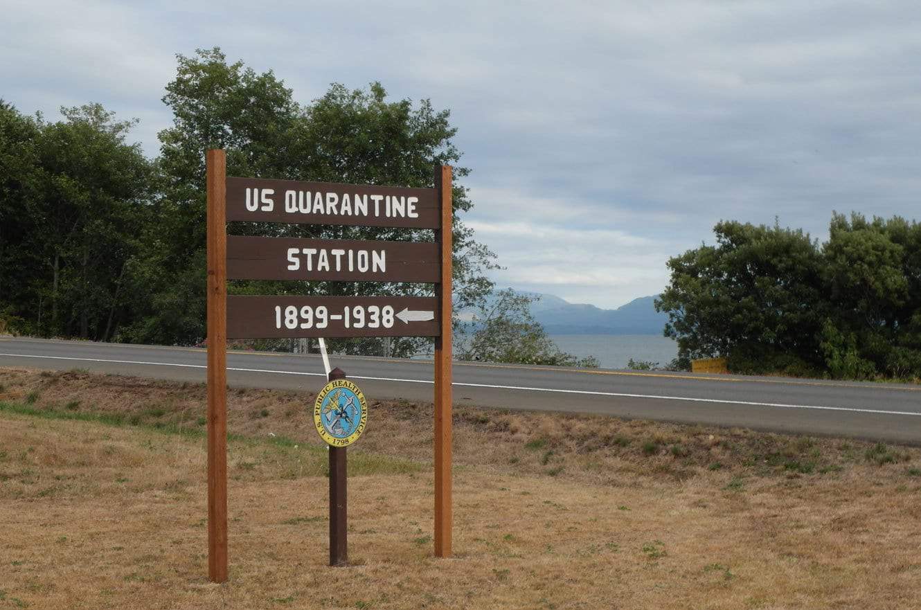 A weathered wooden sign reading "US Quarantine Station 1899-1938" beside the road, with the Columbia River in the background