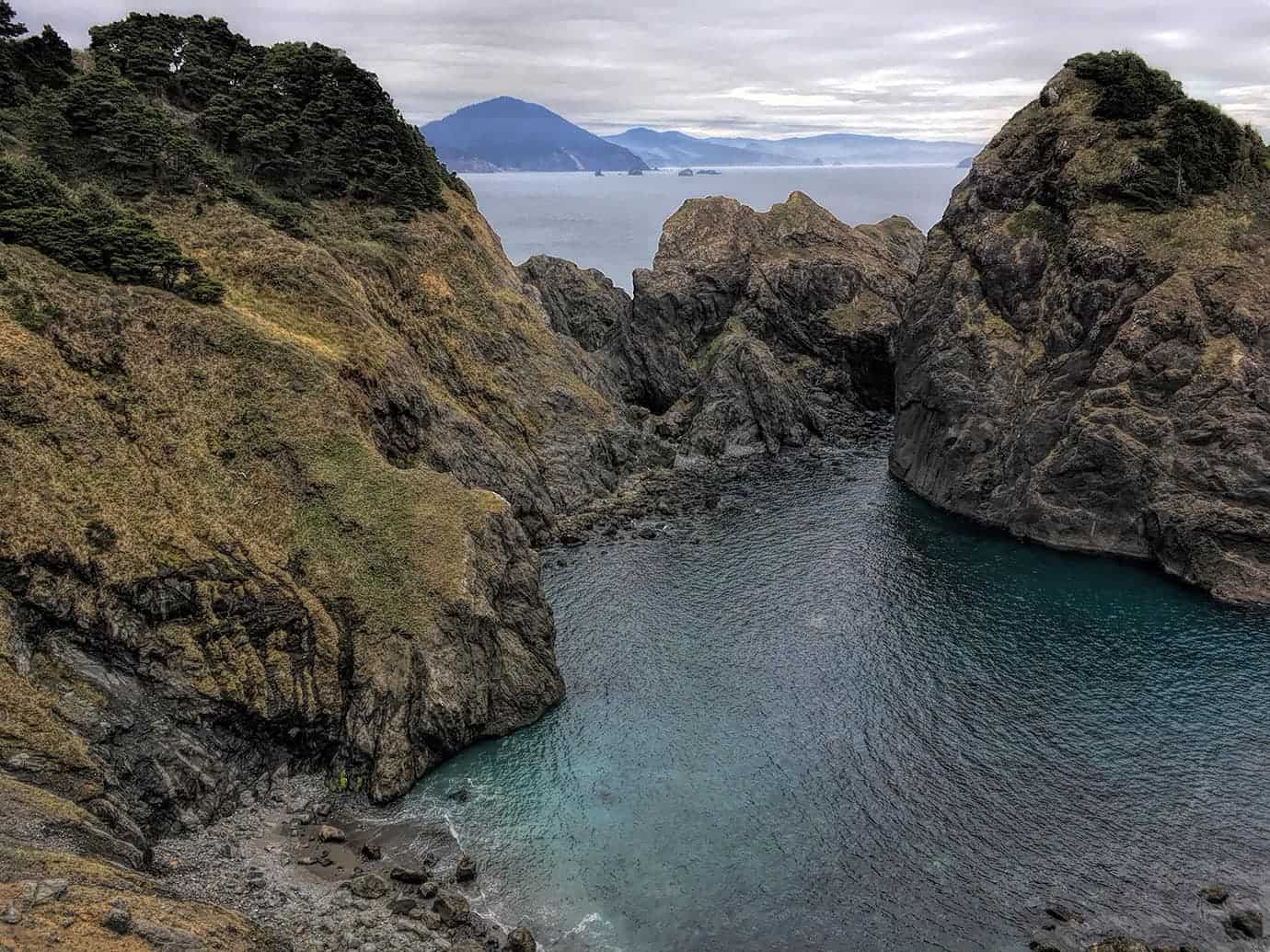 A similar view into the rocky cove between two cliffs, with hazy coastal mountains rising along the horizon beyond the water