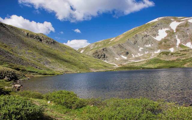 Ptarmigan Lake filling a green alpine basin, framed by a snow-streaked mountain ridge under a partly cloudy sky