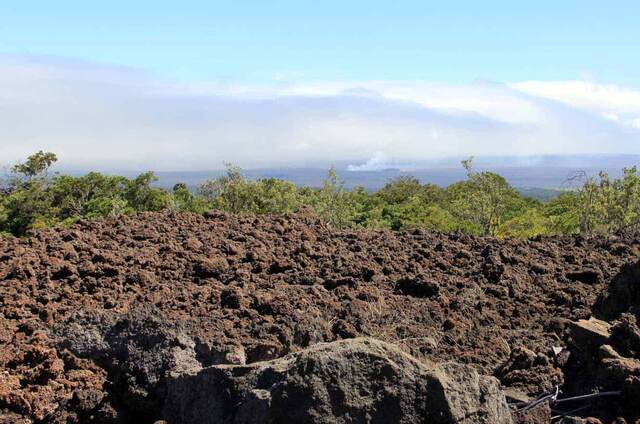 A hazy view over the lava field toward green trees, with smoke rising in the distance