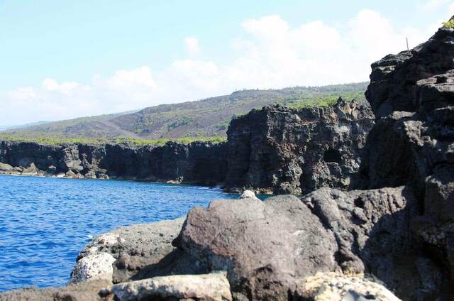 Rugged lava cliffs meeting the ocean along the coastline, with a hillside rising beyond