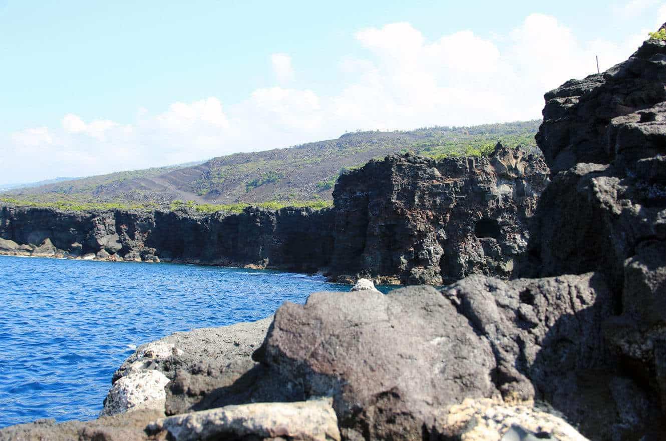 Rugged lava cliffs meeting the ocean along the coastline, with a hillside rising beyond